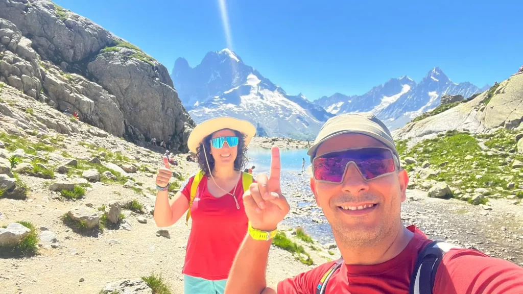 Dos senderistas señalando un lago de alta montaña con picos nevados al fondo.